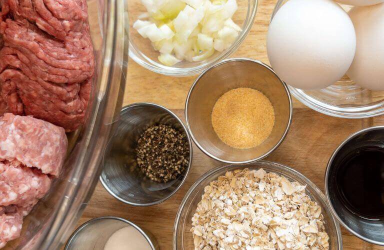 ingredients for a burger recipe classic including ground beef and ground pork in clear bowl with spices, eggs, onions and gluten-free oatmeal