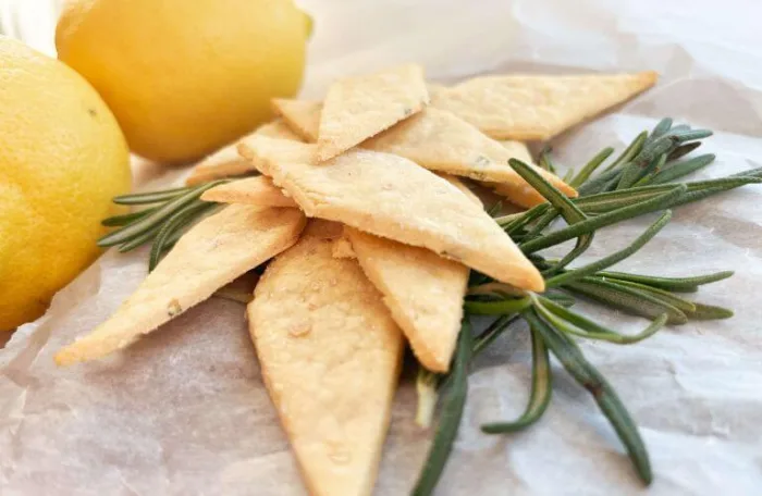 A stack of crispy gluten-free lemon rosemary sourdough discard crackers with a sprig of rosemary and lemons