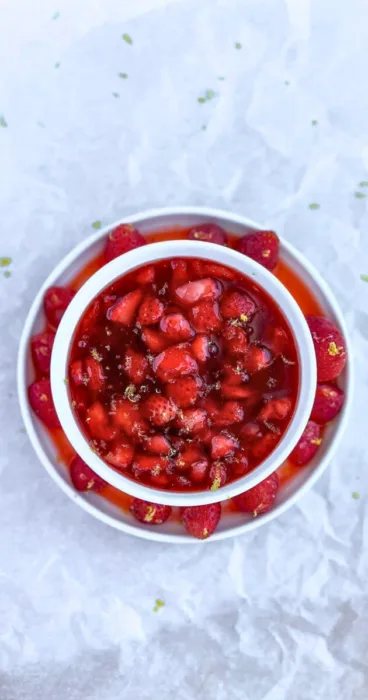 A vibrant display of gluten-free strawberry sauce, with strawberries arranged on a white glass plate against a parchment paper kitchen backdrop.