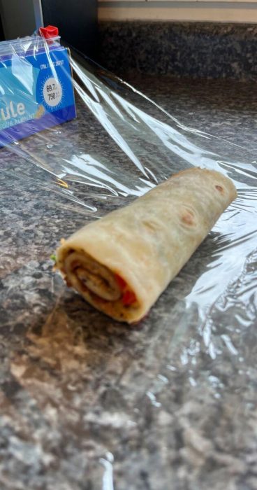 Gluten-free tortilla wrap stuffed with an omelet, vibrant veggies, and bacon wrapped in plastic wrap.